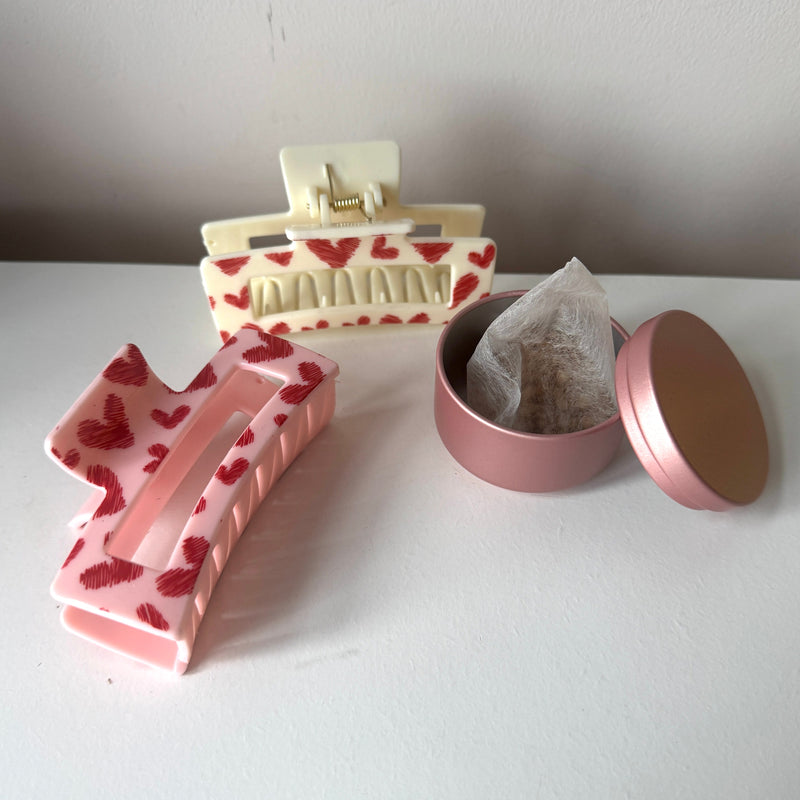 Pink hair clip with heart design and a small pink container with tea bags on a white surface.